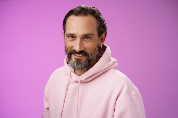 Charming alluring handsome bearded stylish adult male model earring pink hoodie smiling delighted express confidence positivity feel lucky amused, standing purple background talking casually.