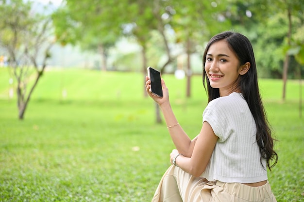 Charming Asian female chilling in the park holding her smartphone sitting on grass