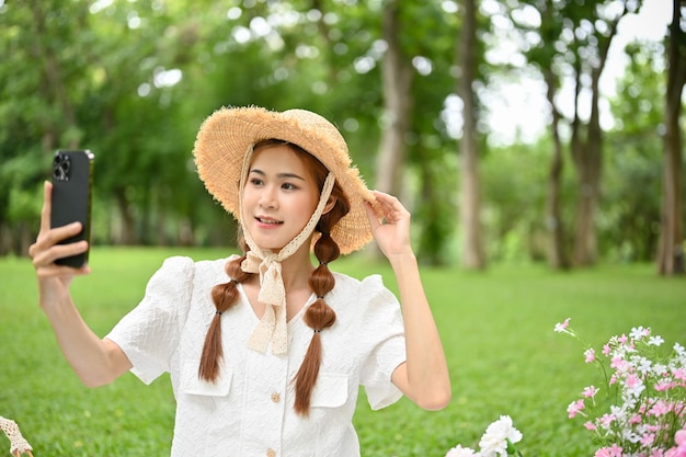 Charming Asian female with a beautiful straw hat strolling in garden and using her smartphone