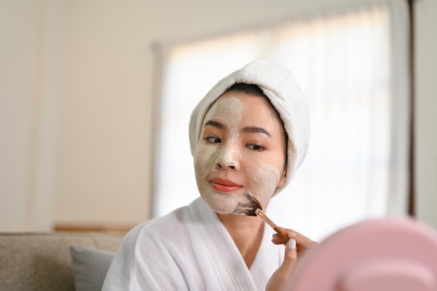 Charming Asian woman in bathrobe looking in the mirror applying detoxifying facial clay mask