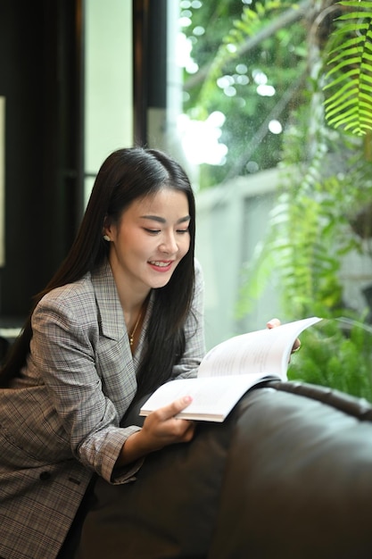 Charming asian woman taking a break from work and reading a book on comfortable couch