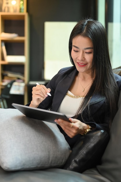 Charming asian woman using digital tablet while resting on sofa in her personal office