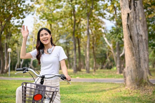 Charming Asian woman waving her hand greeting her friends while she is on her bike in the park