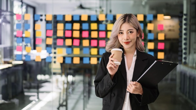 Charming Asian woman with a smile standing holding papers and coffee cup at the office