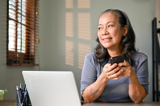 Charming Asianaged businesswoman holding her phone daydreaming at her desk