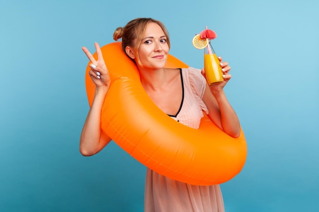 Charming beautiful female with blonde hair and two hair buns holding cocktail and showing victory sign looking at camera with satisfied expression Indoor studio shot isolated on blue background