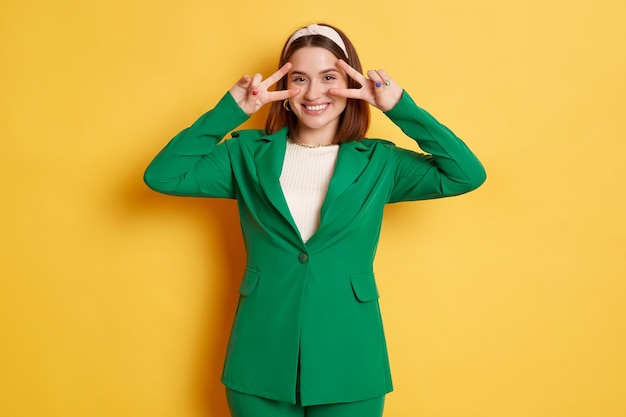 Charming beautiful woman wearing green jacket standing isolated over yellow background showing vi signs near her eyes having fun celebrating expressing happiness