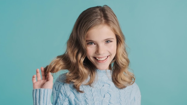 Charming blond teenager girl with wavy hair looking cute smiling on camera isolated on blue background