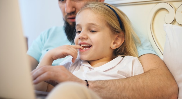 Charming blonde girl spending time with her bearded father lying on the sofa and reading something