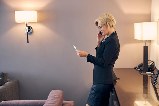 Charming blonde lady in elegant outfit talking on cellphone and holding boarding pass