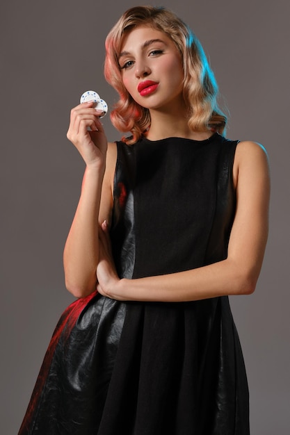 Charming blonde maiden with bright makeup in black stylish dress is holding two chips and posing against gray studio background Gambling entertainment poker casino Closeup