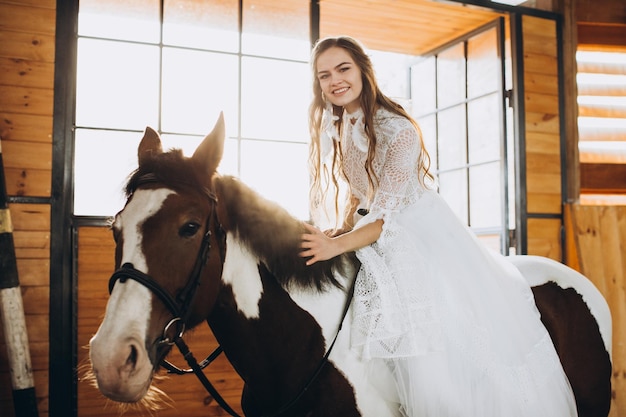 A charming boho bride rides a horse on a ranch at sunset in winter