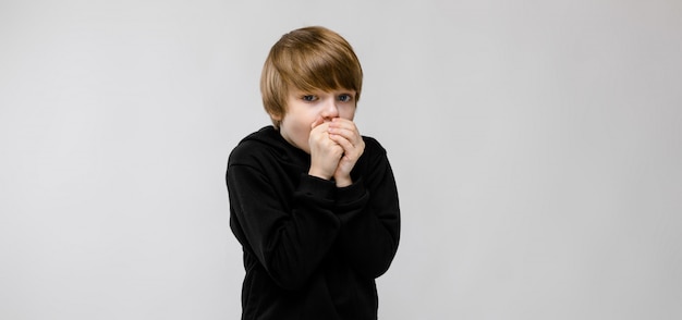 Charming boy with blond hair and dark eyes. The boy raised his hands to his mouth.