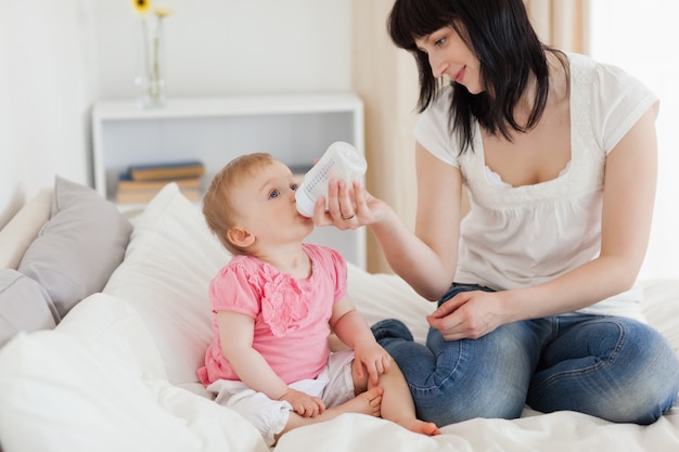 Charming brunette female bottle-feeding her baby on a bed