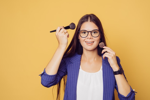 Charming brunette in glasses with long hair dressed in dark blue suit cute smiling, make up herself during quarantine, over yellow background with copy space.