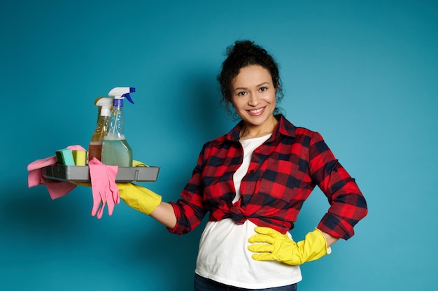 A charming brunette with curly hair in red check shirt and wearing yellow rubber gloves holds cleaning supplies in one hand