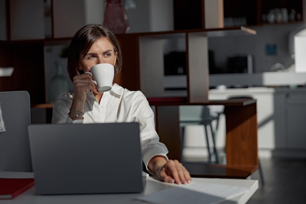 Charming businesswoman working at the office using laptop and drink coffee is looking away