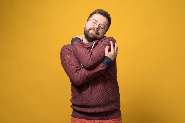 Charming Caucasian bearded man in glasses tenderly hugs himself dreamily closing his eyes