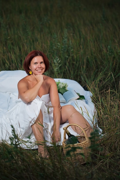 Charming caucasian woman sitting in the white bed in a field at sunset in summer