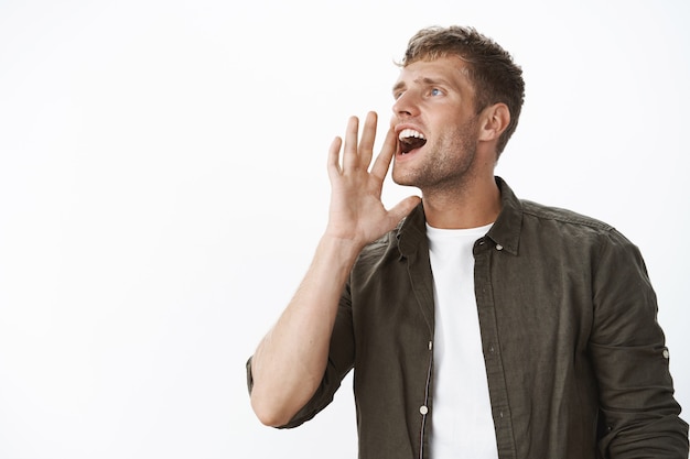 Charming charismatic european blond guy with blue eyes calling someone into distance yelling turning left holding hand near opened mouth as shouting, searching friend in crowd over gray wall