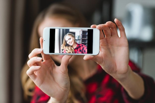 Charming cheerful young woman in plaid shirt taking selfie with smartphone