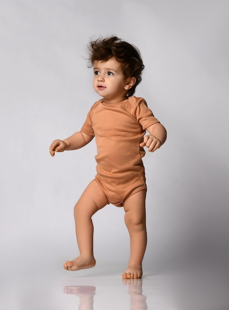 Charming curly baby in beige body is taking its first steps on a gray background