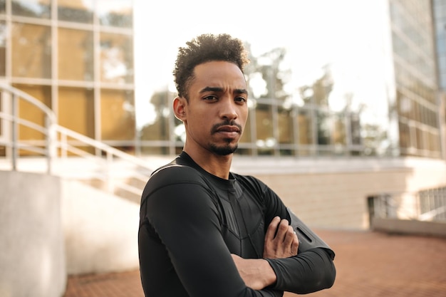 Charming curly brunet man in longsleeved tshirt looks into camera outside Young darkskinned guy in sport outfit crosses arms