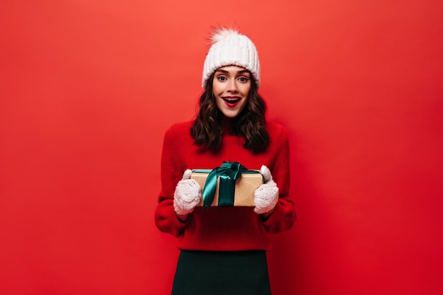 Charming curly girl in red sweater knitted hat and mittens holds gift box and smiles sincerely on red isolated background