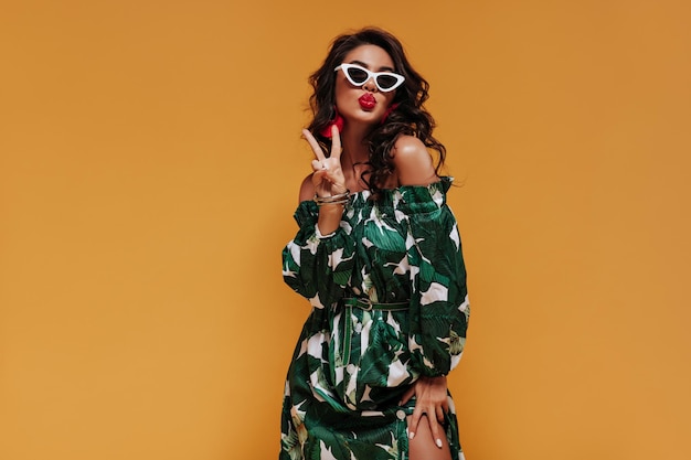 Charming curly haired lady with red lips in green clothes showing peace sign and blowing kiss on orange isolated backdrop