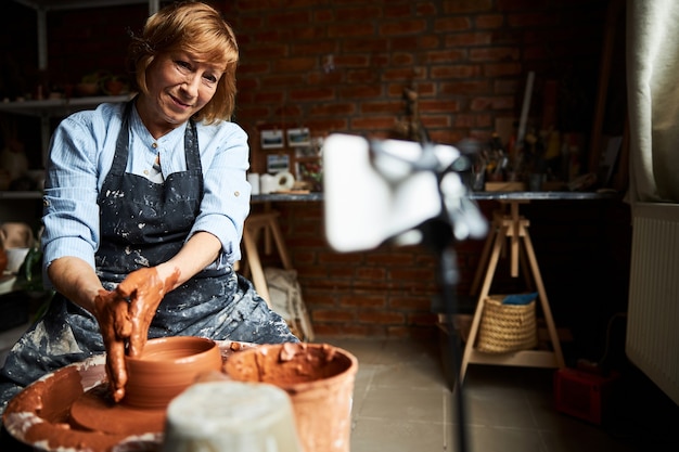 Charming female potter in apron shaping clay and smiling while making pottery