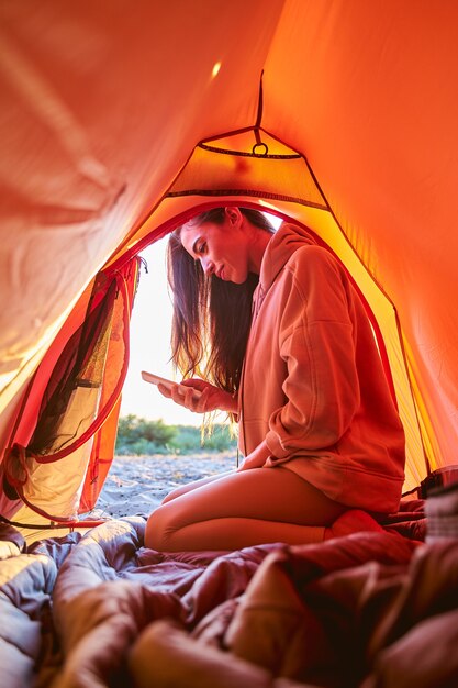 Charming female traveler texting message on smartphone and smiling while sitting in touristic tent