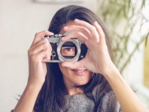 Charming female with long dark hair and brown eyes looking through retro analog photo camera while pretending to taking photo and looking at camera against blurred background