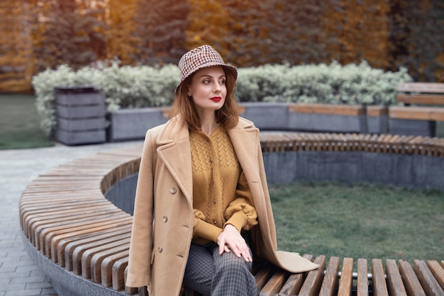 Charming french young woman in an autumn beige coat and plaid hat sits on a round bench flowerbed