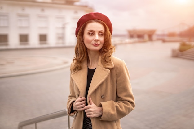 Charming french young woman in an autumn beige coat and red beret standing holding the edges of the