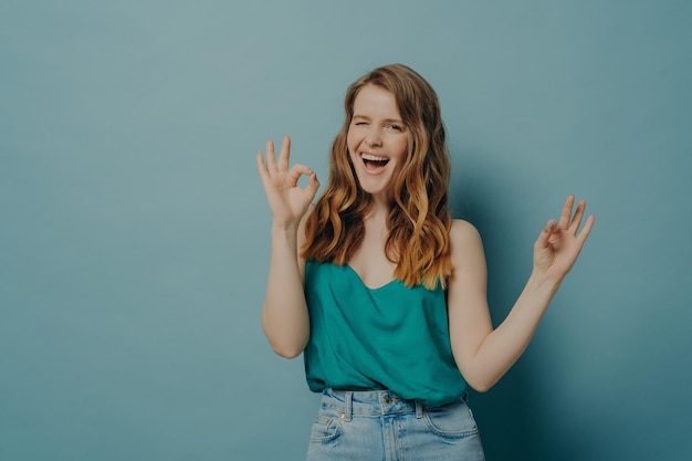 Charming funny girl showing OK sign with both hands while winking and looking at camera with big smile isolated over blue studio background Positive body language and facial expressions concept