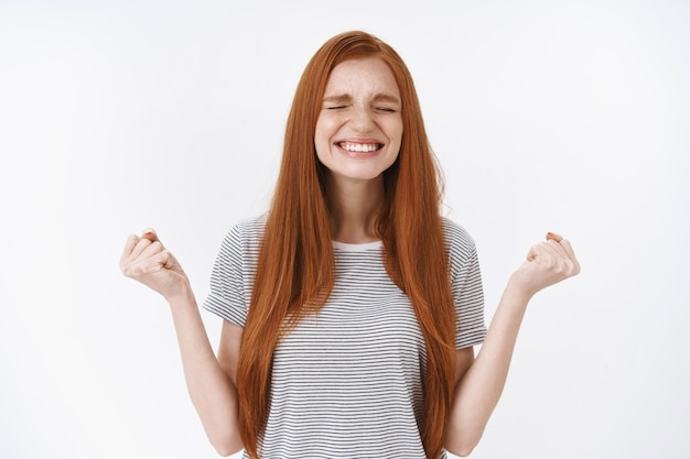 Charming gentle devoted redhead cute girl close eyes smiling delighted clench fists victory triumph gesture achieve goal accomplish good result, standing happily white wall