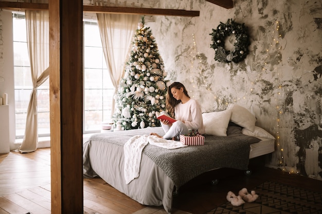 Photo charming girl dressed in white sweater and pants reads a book sitting on the bed with gray blanket, white pillows and a new year gift in a decorated room with a new year tree . .