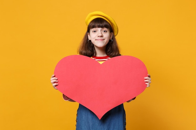 Charming girl teenager in french beret, denim sundress holding big empty blank red heart isolated on yellow wall background in studio. People sincere emotions, lifestyle concept. Mock up copy space.