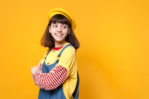 Charming girl teenager in french beret, denim sundress holding hands folded, looking aside isolated on yellow wall background in studio. People sincere emotions, lifestyle concept. Mock up copy space.