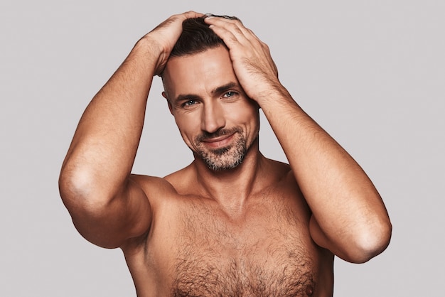 Charming handsome. Charming young man keeping hands in hair and smiling while standing against grey background