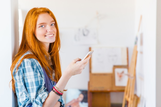 Charming happy young woman with long red hair using smartphone in art workshop