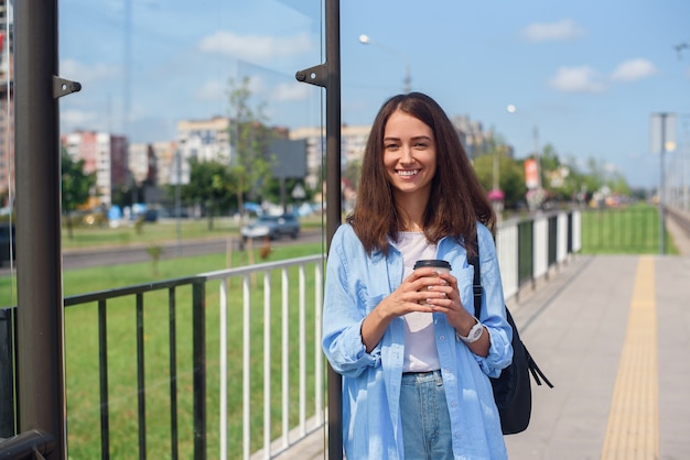 Charming hipster girl waiting for bus or tram on public transport station in the morning with cup of coffee