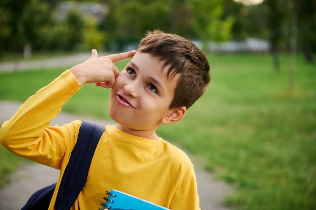 A charming joyful cheerful schoolboy holds his hand to his temple imitating a pistol, as a sign of tiredness from studying after a hard day at school, looking up in the city park background