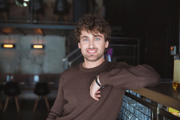 Charming joyful content handsome positive charismatic young man in brown sweetshirt posing in cafe