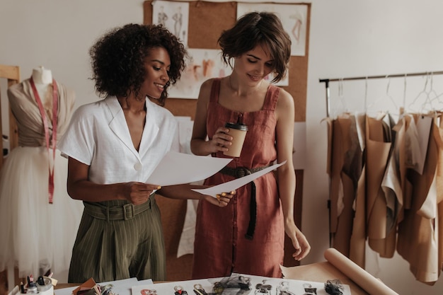 Charming ladies look at paper sheets Curly darkskinned woman in white blouse and shorthaired lady in linen dress work as fashion designers