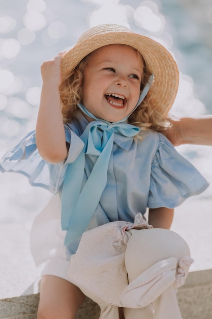 charming little blond girl in a straw hat and a blue dress plays with a stuffed rabbit outdoors