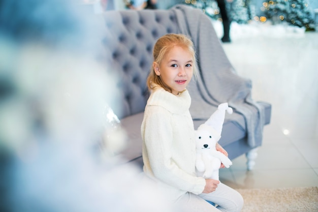 Charming little blonde girl in white warm sweater with toy bear sitting on the blue armchair