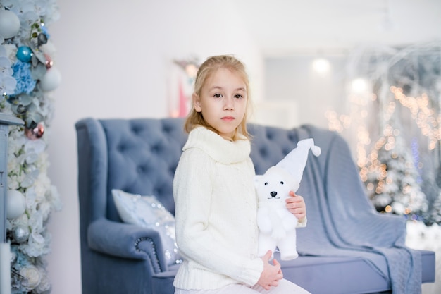 Charming little blonde girl in white warm sweater with toy bear sitting on the blue armchair