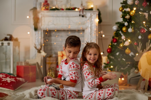 Charming little girl and boy in pyjamas sitting on plaid with presents and christmas tree near blowi...