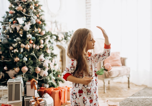 Charming little girl dressed in pajama holds a flower in the full of light cozy room with the New Year's tree .